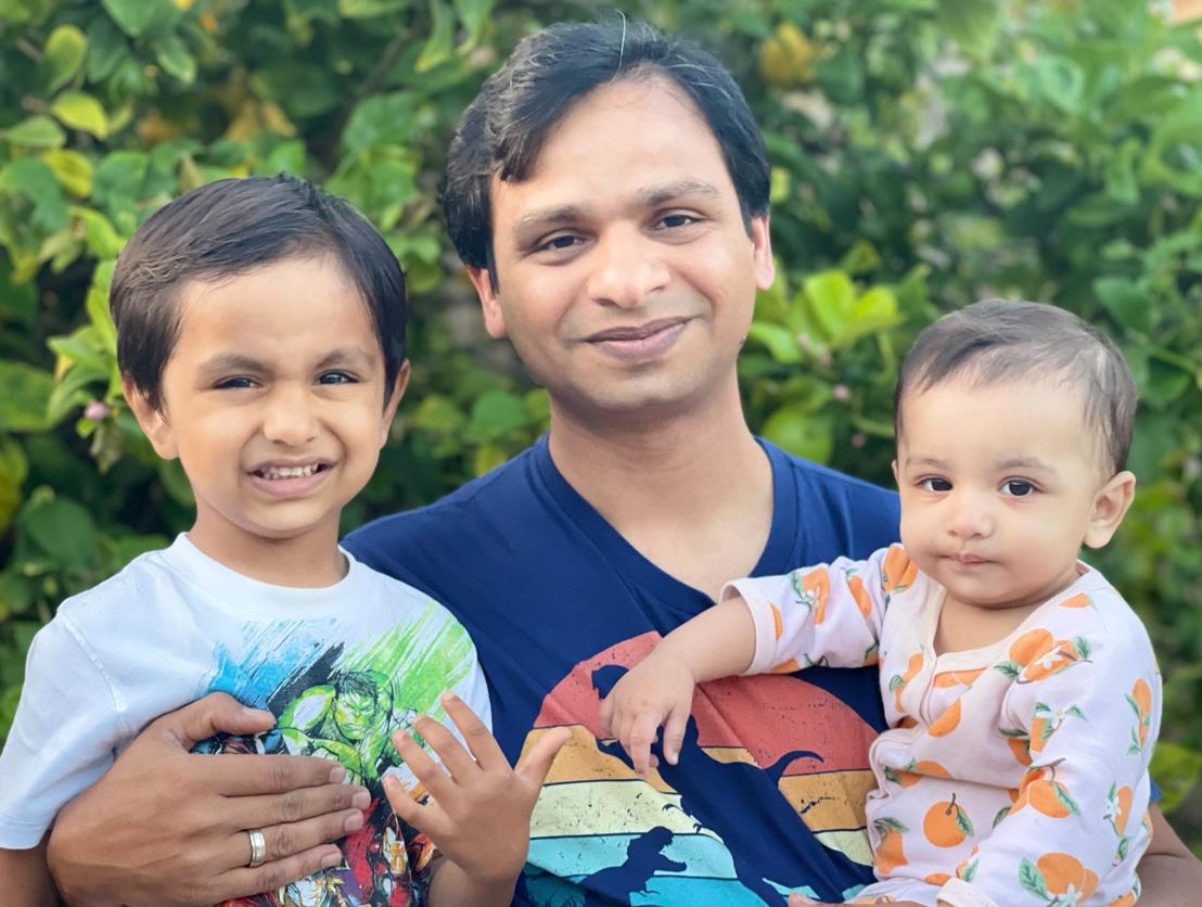 AWS SDE Mahesh Akula holds his two children against a backdrop of foliage and smiles at the camera.